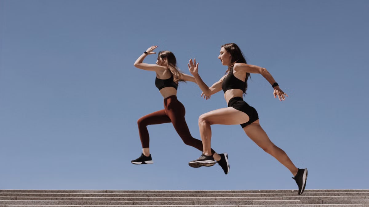 Two women in active wear leap energetically on stairs against a clear blue sky.